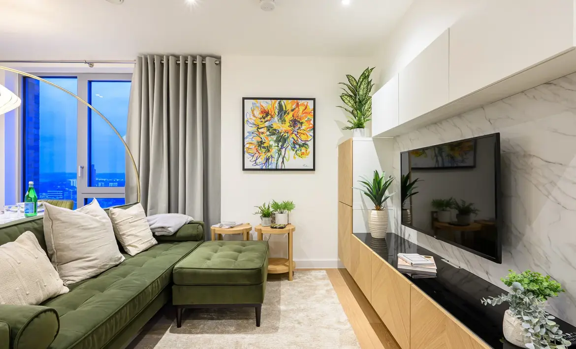 Bright living room featuring a green corner sofa, wall-mounted television, built-in storage units, large window and modern décor.